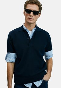Mann trägt dunkle Sonnenbrille, einen marineblauen Pullover über einem blauen gestreiften Hemd mit hochgekrempelten Ärmeln und hat seine linke Hand in der Jeans-Tasche.