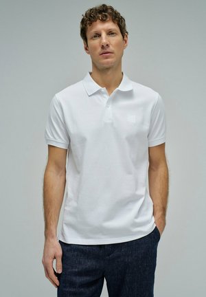 Homme aux cheveux bouclés portant un polo blanc uni à manches courtes et un pantalon foncé, debout devant un fond gris neutre.