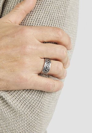 Männliche Hand mit geflochtenem Silberring, die auf einem beigen Strickstoff ruht, die Finger leicht um den Arm gekrümmt.