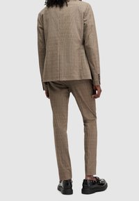 Hnědý kostkovaný oblek se strukturovanou bundou a slim-fit kalhotami. Výrazná textura a elegantní černé boty dotvářejí celkový vzhled.