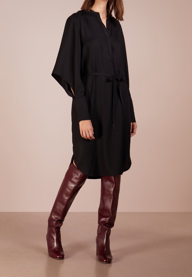 Femme portant une robe noire à manches longues avec une ceinture à la taille et des bottes en cuir bordeaux montantes jusqu'aux genoux, debout sur un sol beige.