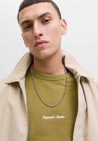 Giacca beige con zip indossata sopra un felpa verde con collo a coste e testo ricamato bianco. Collana a catena argentata visibile.
