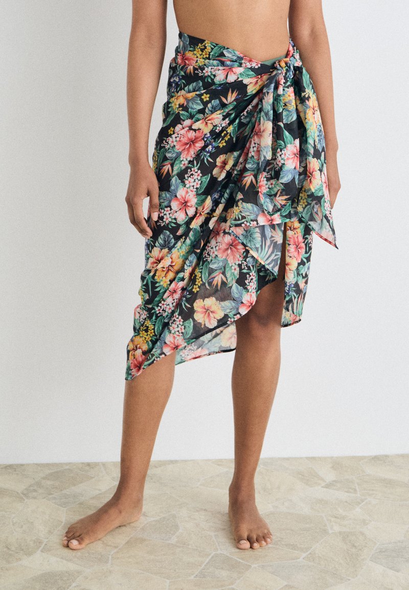 Sarong bermotif bunga dengan warna-warna cerah, kain ringan, sedikit transparan, hem asimetris, dan detail pinggang yang diikat.