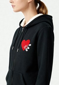 Zwarte hoodie met een rood hartontwerp en witte veiligheidsspelden, met een gestructureerde fleece voering en zilveren ritsaccenten.
