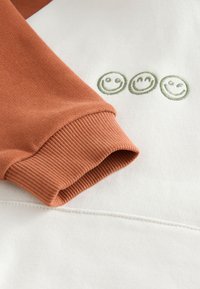 Mikina z bavlneného mixu s oranžovým rukávom a bielym telom, ozdobená zelenými vyšívanými usmievavými tvárami pri leme a rebrovanými manžetami.