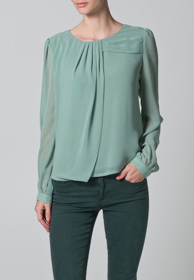 Blouse vert menthe avec un devant plissé, des manches longues en voile transparent et un col rond. Comprend une petite poche sur le côté gauche de la poitrine et des manches à poignets.