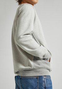 Felpa grigia realizzata in tessuto morbido, con maniche lunghe, polsini a costine, tasche laterali e una vestibilità rilassata. Indossata con jeans blu.