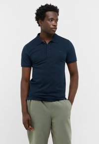 Granatowa koszulka polo z kołnierzykiem i krótkimi rękawami, z subtelnym logo na piersi. Gładka faktura, w połączeniu z jasnoszarymi spodniami.