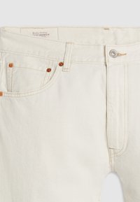 Jean en denim blanc cassé avec rivets en cuivre, poches avant, fermeture par boutons et passants de ceinture, visible de la taille à la partie supérieure de la cuisse.