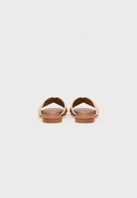 Flate sandaler med to vevde stropper i en lys tan-farge, avrundet form, og en brun flat såle, uten synlig maskinvare eller detaljer.