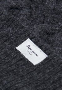 Texture di un maglione lavorato a maglia grigio scuro con un aspetto peloso. Presenta un'etichetta in tessuto bianco con la scritta "Pepe Jeans London" in testo nero.