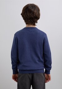 Suéter de punto azul marino con mangas largas y cuello redondo. Textura suave, ajustado en la cintura, usado sobre pantalones de pana gris.