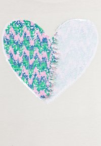 Hvitt stoff har et stort, teksturert hjerte-design. Den ene siden er dekket av paljetter i rosa, blå og grønne sikksakk-mønstre; den andre siden er glatt og pastellfarget.