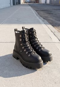 Paire de bottines à plateforme en cuir noir avec lacets et semelles épaisses et robustes posées sur un trottoir en béton ensoleillé.