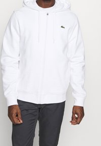 Witte zip-up hoodie van zacht stof, met een klein groen Lacoste-logo, treklinten en geribbelde manchetten, gecombineerd met donkergrijze broek.