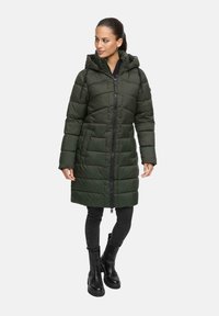 Vrouw in een lange donkergroene gewatteerde pufferjas met capuchon, zwarte jeans en zwarte enkellaarsjes, staande tegen een witte achtergrond.