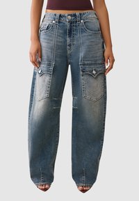 Luźne niebieskie dżinsy z wyraźnymi przeszyciami i dużymi przednimi kieszeniami, o wyblakłym kolorze i przetartym wyglądzie.