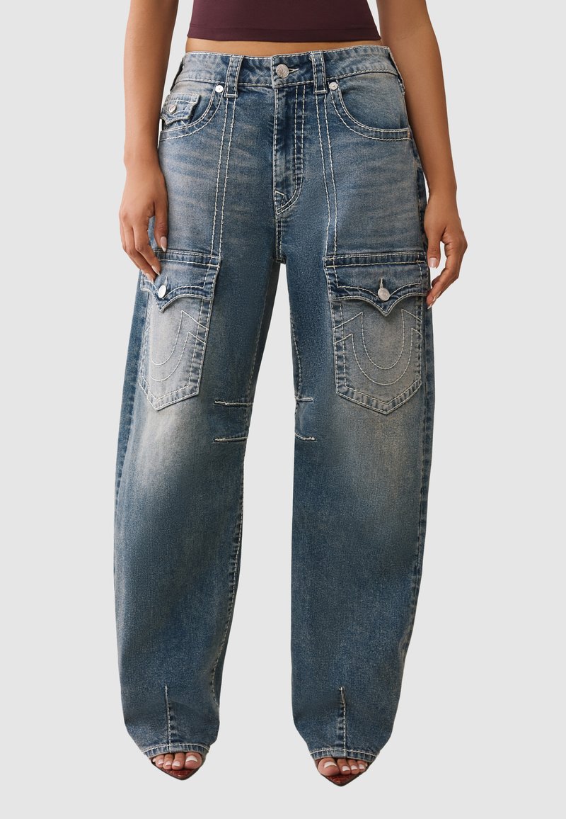 Luźne niebieskie dżinsy z wyraźnymi przeszyciami i dużymi przednimi kieszeniami, o wyblakłym kolorze i przetartym wyglądzie.