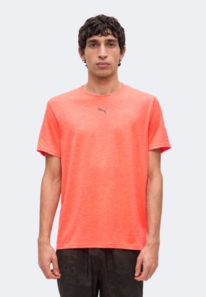 Mann mit kurzen dunklen Haaren, der ein leuchtend oranges Puma-T-Shirt und dunkle Hosen trägt, steht vor einem einfarbigen hellen Hintergrund.