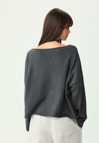 Dunkelgrauer, übergroßer Pullover mit weitem Ausschnitt und überschnittenen Schultern; strukturierter Stoff; kombiniert mit hellgrauen Hosen.
