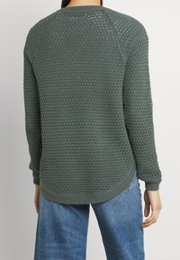 Grüner, strukturierter Pullover mit lockerem Strickdesign, rundem Ausschnitt und gerippten Bündchen. Kombiniert mit blauen Jeans aus Denim mit abgerundetem Saum.