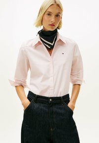 Camisa de botones a rayas rosa claro con mangas remangadas, vaqueros negros de tiro alto y pañuelo de cuello con estampado en blanco y negro.