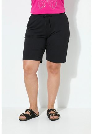Persoon draagt zwarte knielange shorts met trekkoord en zwarte instapsandalen met gespen en studs, staand tegen een witte achtergrond.
