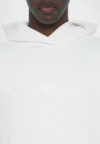 White hoodie featuring an embossed logo reading "QUARTZ CO" on the front. The material appears soft with a smooth texture and a relaxed fit.