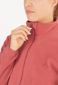 Fleecejakke i støvet rosa med høj krave og lynlås. Har broderet logo og glat tekstur. Lynlåsuddtrækkeren er sort.