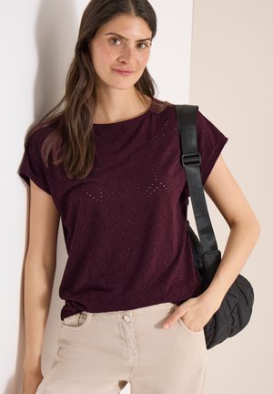 Kvinde iført en mørkelilla, perforeret kortærmet top, lysebeige bukser og en sort, quiltet skuldertaske.