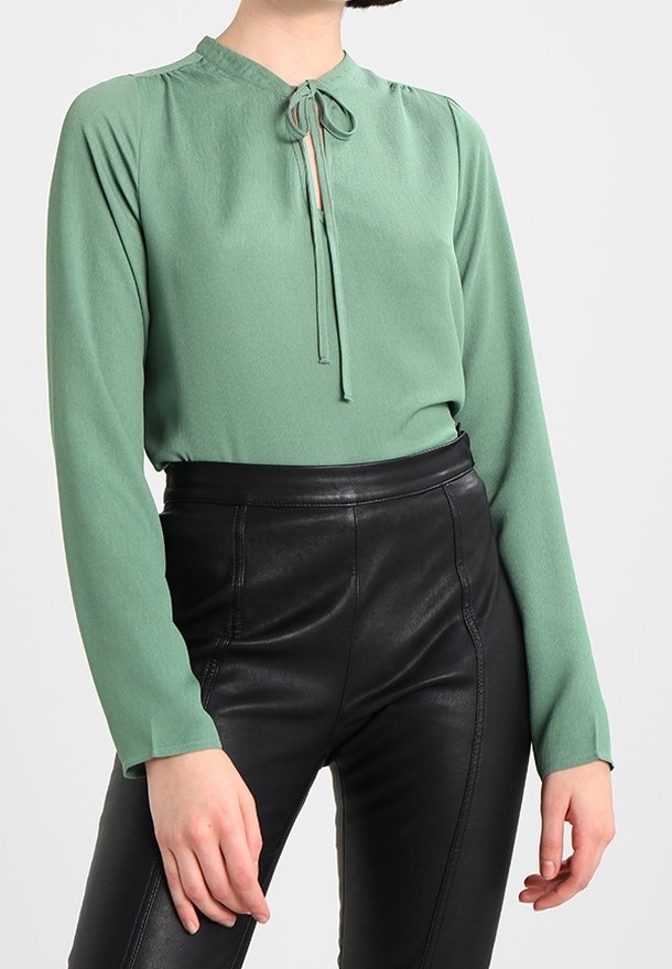 Femme portant un chemisier vert à manches longues avec un col à nœud papillon et un pantalon en cuir noir taille haute sur un fond uni.