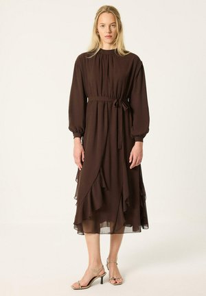 Femme blonde debout portant une robe midi marron foncé à manches longues avec une ceinture à la taille et une jupe à volants superposés, associée à des talons beiges à brides.