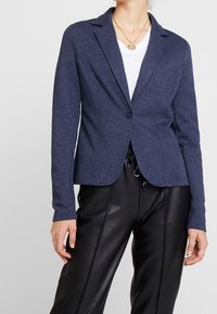 Blazer azul marino de tela suave con cierre de un solo botón, corte entallado, solapas de pico y mangas largas, combinado con pantalones de cuero negro.