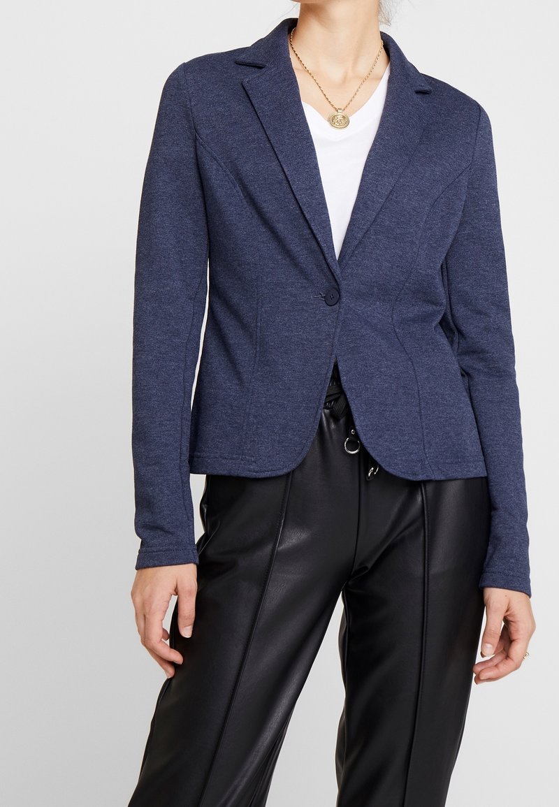 Blazer azul marino de tela suave con cierre de un solo botón, corte entallado, solapas de pico y mangas largas, combinado con pantalones de cuero negro.