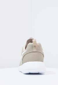 Beige nät-sneaker med vit sula, visad bakifrån, med en dragflik på hälen och en vit swoosh-design på sidan.