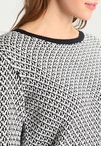 Femme portant un pull en maille noir et blanc avec un petit motif pied-de-poule et un col rond noir, montrant le cou et la zone des épaules.