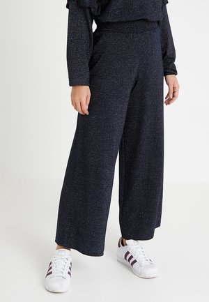 Persoon die donkere wijde broek en witte Adidas-sneakers met paarse strepen draagt, staand tegen een effen lichte achtergrond.