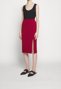 Femme portant un haut noir sans manches avec des bords festonnés, une jupe rouge longueur genou avec une fente latérale, et des chaussures noires pointues sans lacets.
