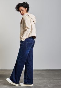 Vaaleanbeige lyhyt farkkutakki hupulla, yhdistetty pimeisiin sinisiin leveälahkeisiin farkkuihin. Valkoisia lenkkareita kengissä, kääntyen hieman taaksepäin.