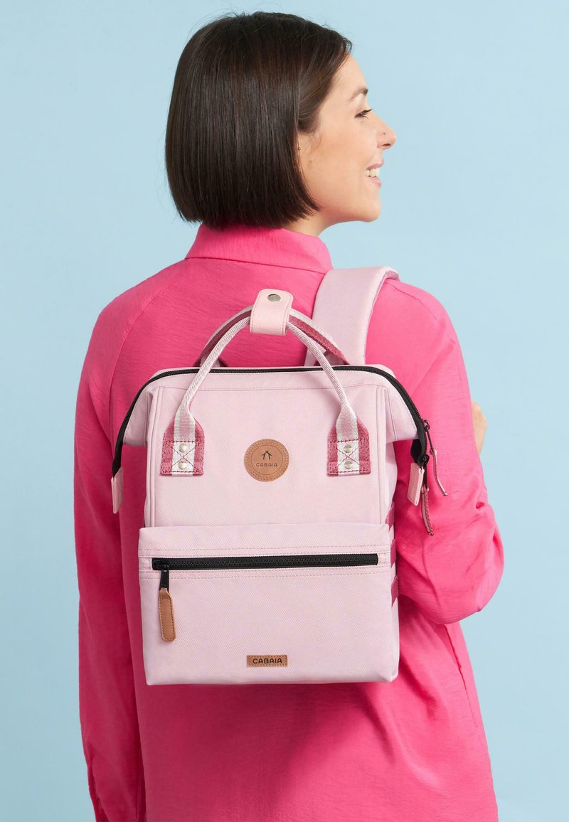 Hellrosa Rucksack aus strukturiertem Material, mit zwei oberen Griffen, seitlichen Reißverschlüssen und einer Fronttasche. Braunes Markenlogo und Akzente in dunklerem Pink.