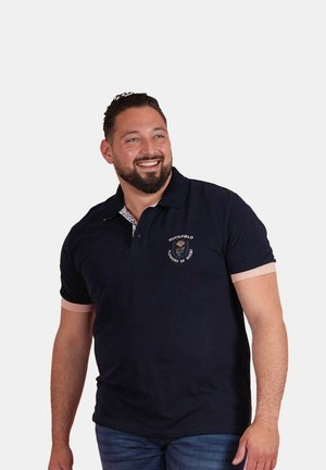 Homme souriant aux cheveux courts et foncés et à la barbe, portant un polo bleu marine avec le logo « Ruckfield Powers of Rugby » et un jean bleu, sur un fond uni.