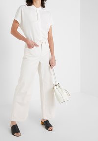 Žena v bílých širokých kalhotách a bílém tričku s krátkým rukávem, která drží bílou kabelku a má na sobě černé sandály na podpatku s otevřenou špičkou.