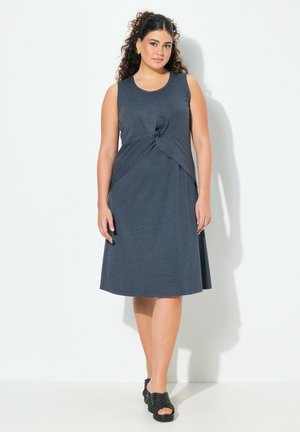Ärmelloses dunkelblaues Kleid mit einem geknoteten Frontdetail, fließendes A-Linien-Design, aus weichem Stoff. Kombiniert mit schwarzen strukturierten Sandalen.