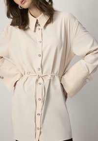 Button-up shirt in light beige, featuring a collar, long sleeves with wide cuffs, a tie belt at the waist, and silver-tone buttons.