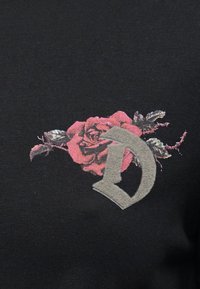 Ροζ τριαντάφυλλο με πράσινα φύλλα, μερικώς καλυμμένο από ένα υφασμάτινο γκρι γράμμα "Δ" σε μαύρο ύφασμα.