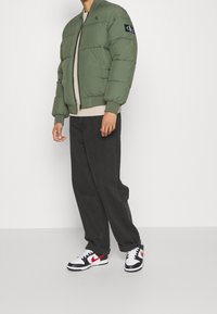 Groene pufferjas met ritssluiting en een zwarte logopatch, gecombineerd met grijze broek en zwarte en rode sneakers. Effen achtergrond.