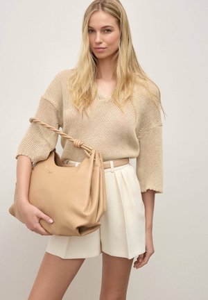 Donna bionda con maglione beige in maglia e pantaloncini bianchi che tiene una grande borsa di pelle color cuoio con manico intrecciato su sfondo neutro.