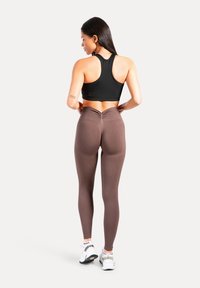 Brassière de sport noire et leggings marron taille haute avec un détail froncé dans le dos. Modèle portant des baskets blanches, debout sur un fond uni.