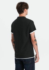 Svart polo skjorta med vit kant på ärmar och nederkant, i bomullsmaterial, kortärmad och med en klassisk krage. Bakvy visas.