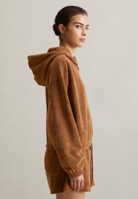 Hoodie en polaire marron avec corde de serrage ; coupe ample et poche kangourou ; porté avec un short assorti ; la vue de côté met en valeur la texture et la forme.